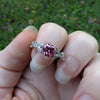 Heart-shaped pink gemstone ring on a finger with a green outdoor background