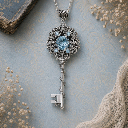 Silver key-shaped pendant with blue gemstone on a textured surface.  Aquamarine Fantasy Key Necklace – Elven Fairycore Swarovski Crystal Jewelry