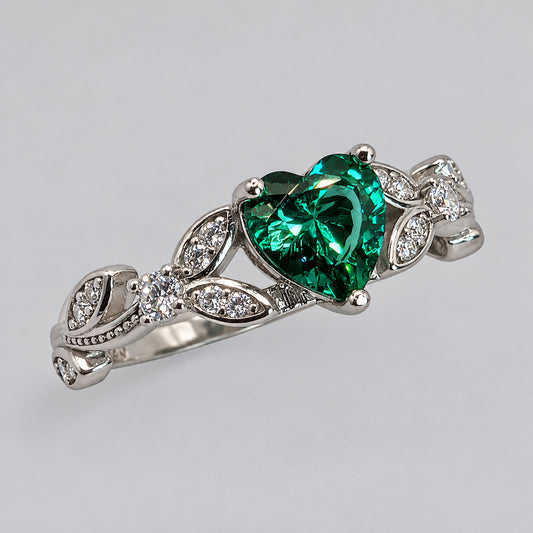 A sterling silver ring featuring a heart-shaped green emerald cubic zirconia center stone and small clear stones set along leaf-like side motifs, photographed on a light gray background.