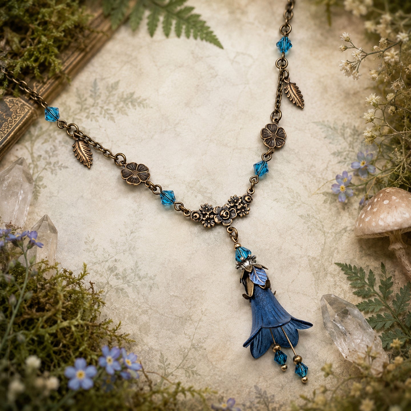 An ornate woodland-style necklace featuring a blue bell-shaped flower pendant with gold detailing, accented by blue faceted Swarovski crystal beads and antique brass filigree charms, displayed on a textured parchment background surrounded by moss, crystals, and small flowers.