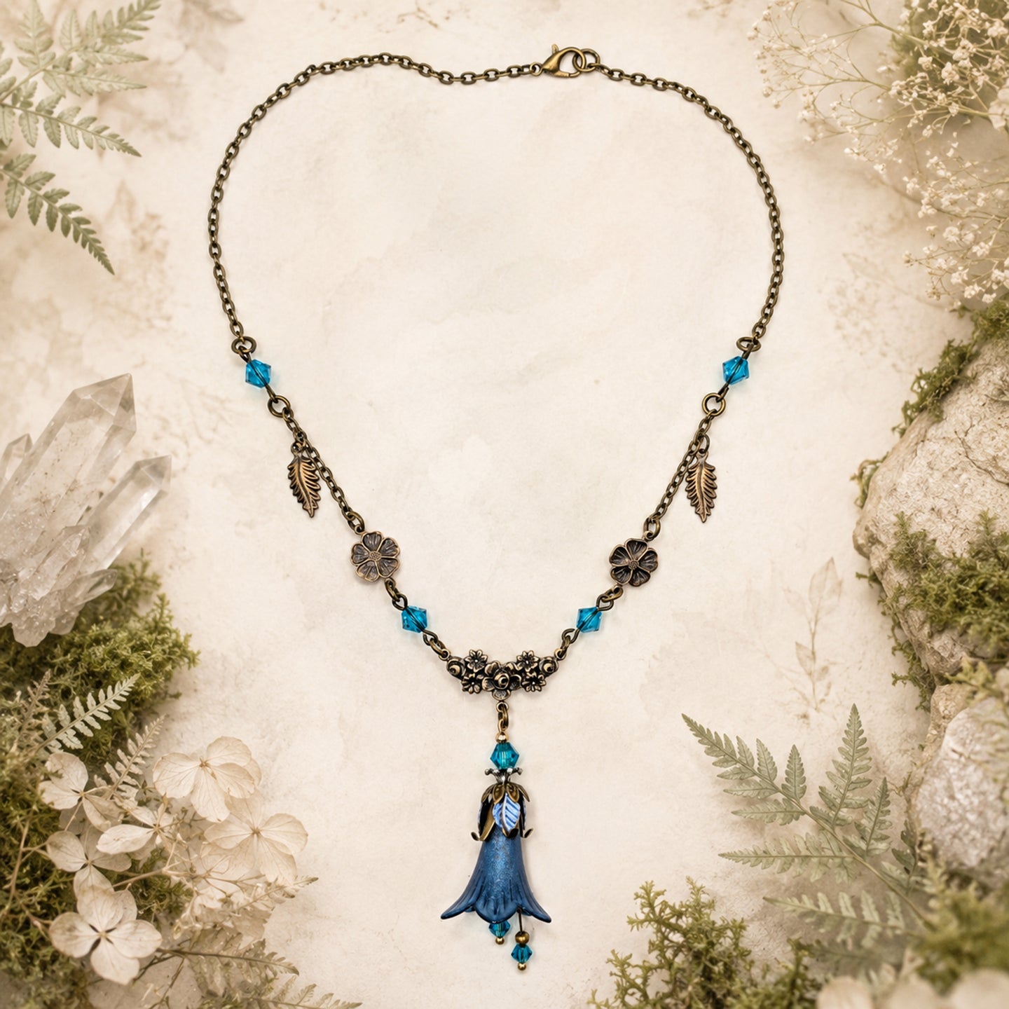 An ornate woodland-style necklace featuring a blue bell-shaped flower pendant with gold detailing, accented by blue faceted Swarovski crystal beads and antique brass filigree charms, displayed on a textured parchment background surrounded by moss, crystals, and small flowers.