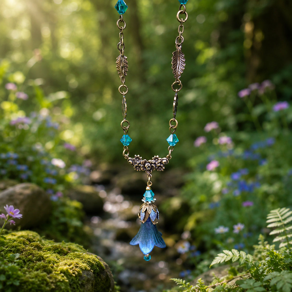 A fairy nature-styled floral necklace with turquoise blue crystal beads, a central flower filigree cluster, and a dangling blue bell-shaped pendant, photographed hanging against a blurred forest stream background.