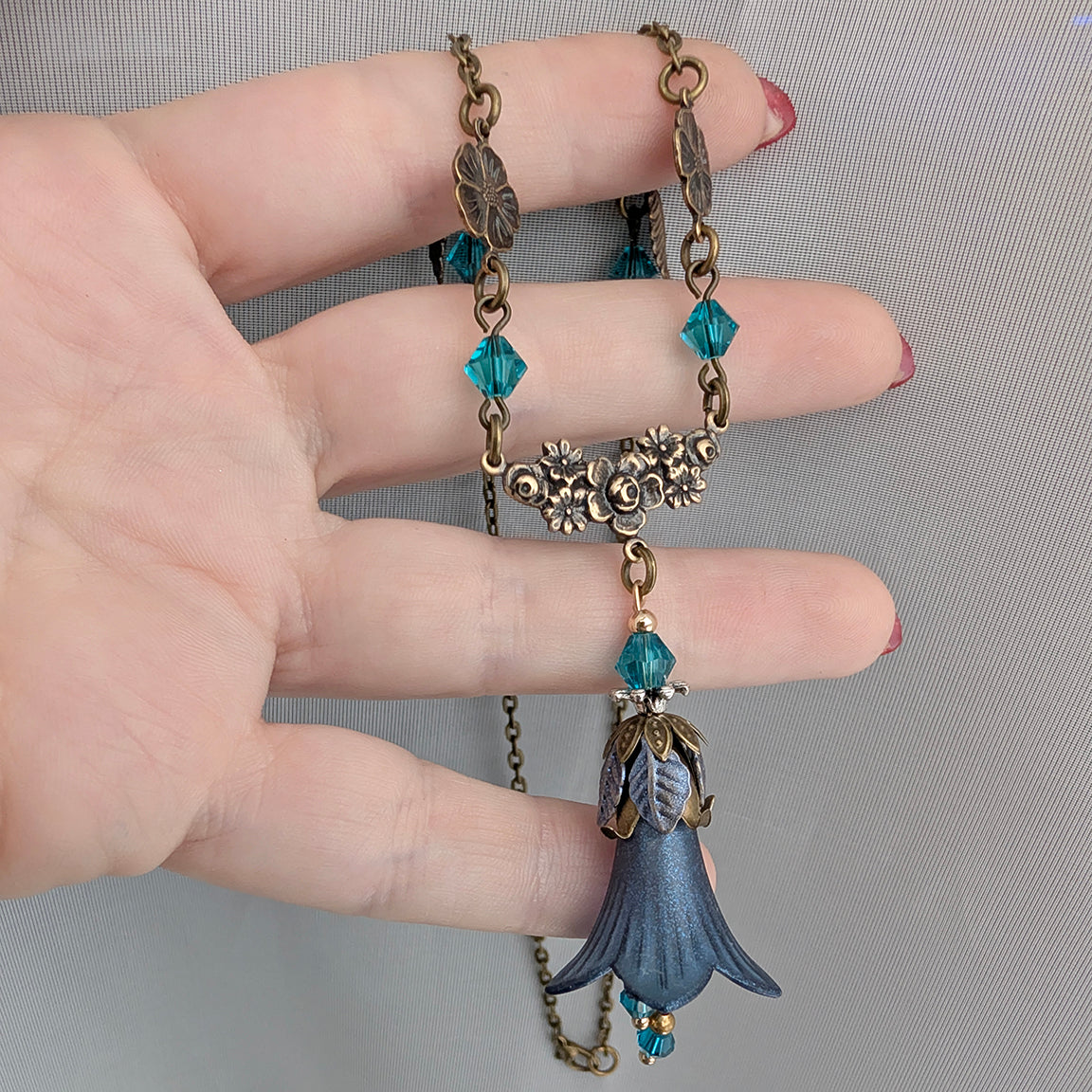 A close-up of a hand holding a vintage-style fairy woodland floral necklace with antique brass filigree flowers, teal faceted crystal beads, and a blue bell-shaped pendant.