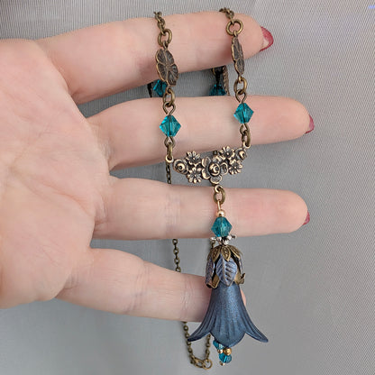 A close-up of a hand holding a vintage-style fairy woodland floral necklace with antique brass filigree flowers, teal faceted crystal beads, and a blue bell-shaped pendant.