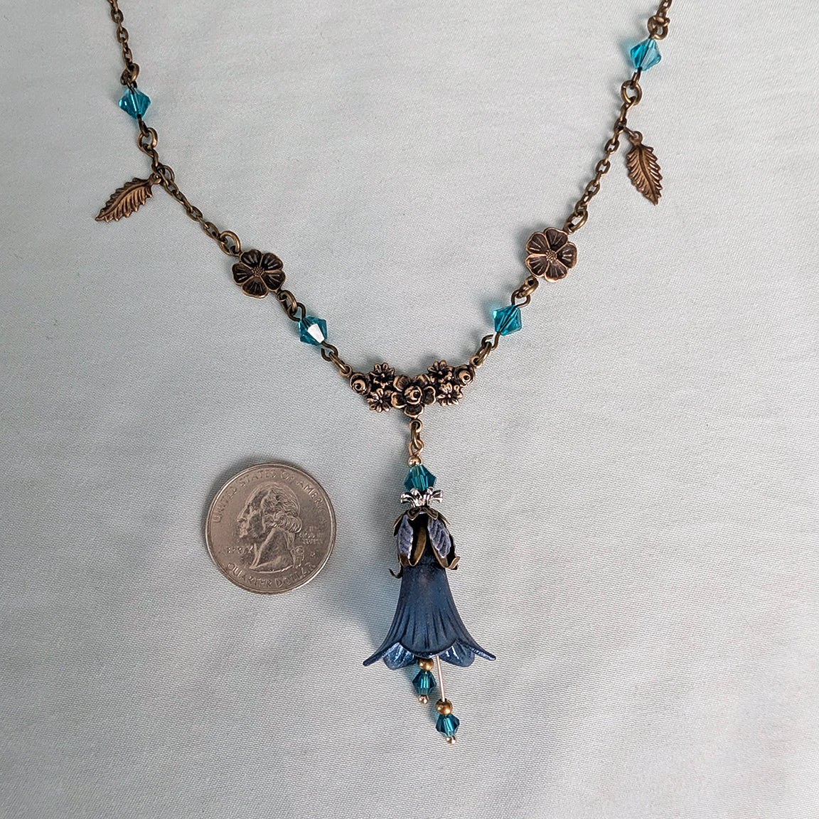 An antique-style bronze chain floral nature-inspired necklace with blue faceted crystal beads, small leaf charms, and a central blue bell-shaped flower pendant, shown on a white fabric background next to a US quarter coin for scale.