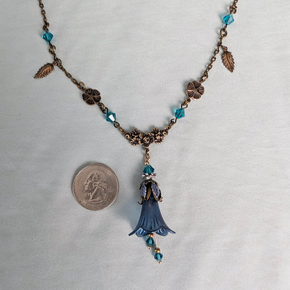 An antique-style bronze chain floral nature-inspired necklace with blue faceted crystal beads, small leaf charms, and a central blue bell-shaped flower pendant, shown on a white fabric background next to a US quarter coin for scale.