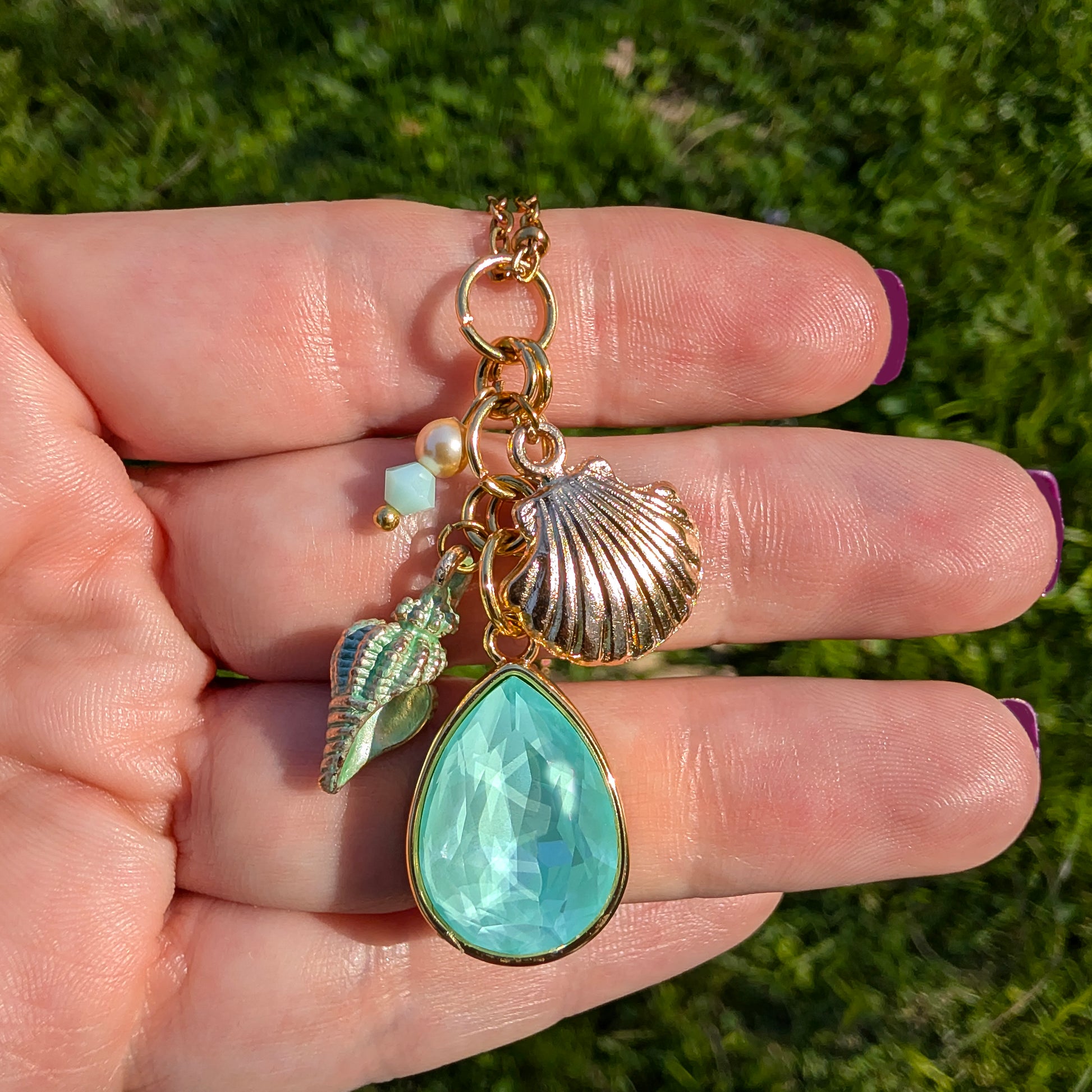 Hand holding a gold charm with a green teardrop pendant, pearl, and shell against a grass background