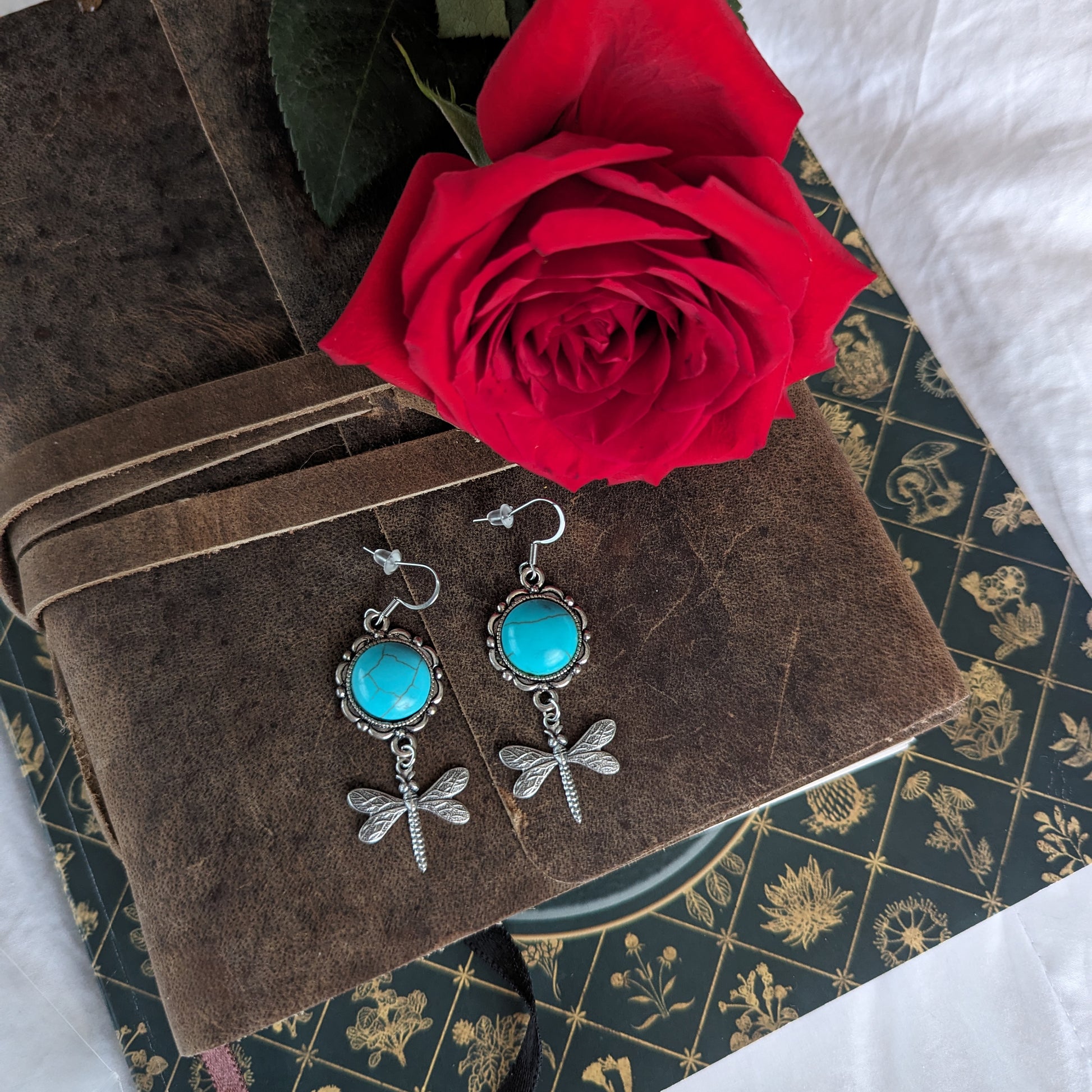 Dragonfly earrings with turquoise stones on a brown fabric background with a red rose.