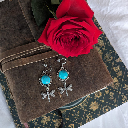 Dragonfly earrings with turquoise stones on a brown fabric background with a red rose.