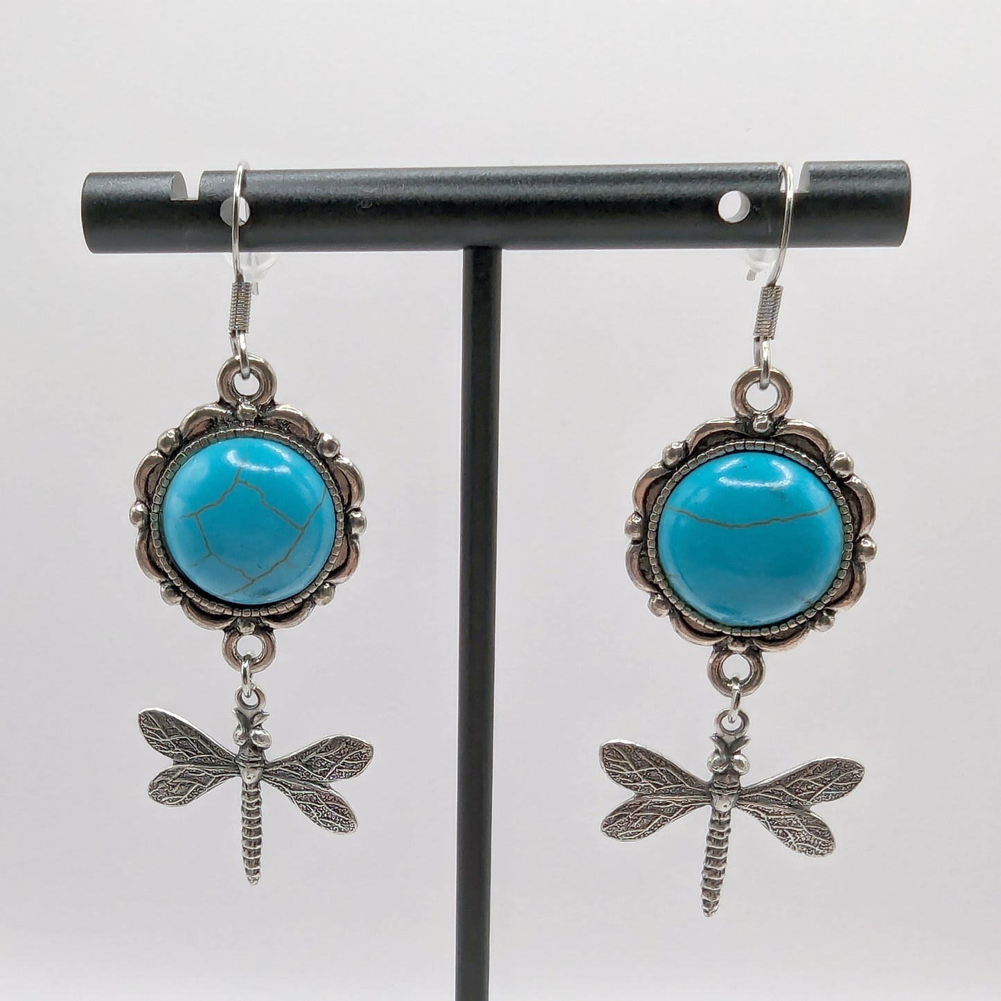 Turquoise stone earrings with dragonfly charms on a black stand against a white background