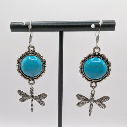 Turquoise stone earrings with dragonfly charms on a black stand against a white background