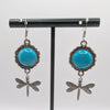 Turquoise stone earrings with dragonfly charms on a black stand against a white background