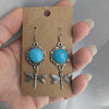 Turquoise earrings with silver dragonfly designs on a brown card held by a hand.
