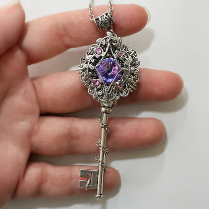 Decorative key-shaped pendant with purple gemstone held by a hand on a white background.  Fairycore Silver Key Necklace with Swarovski Crystal – Fantasy Floral Jewelry