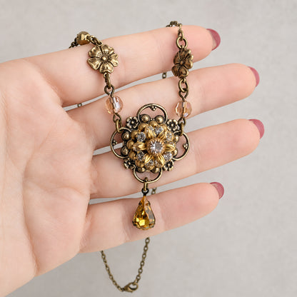 An antique-style brass sunflower shaped pendant necklace with a topaz yellow teardrop crystal drop and small crystal beads, held in a hand with pink nail polish against a neutral gray background.