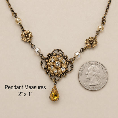 An antique-style flower necklace with a central floral sunflower medallion with crystals, small gold beads, honey faceted beads, and a teardrop-shaped light topaz crystal pendant, displayed on a textured cream background with a quarter for size comparison.