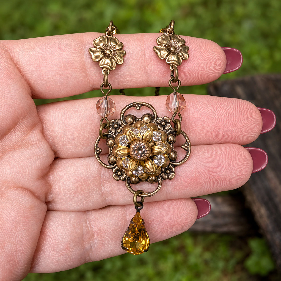 An antique-style floral brass sunflower shaped pendant with clear crystala and a teardrop-shaped amber gemstone held in a hand with pink nail polish against a neutral grassy background. 