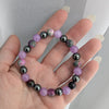 Hand holding a bracelet with purple and black beads against a neutral background