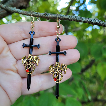 Sword and dragon earrings held between fingers with a natural background