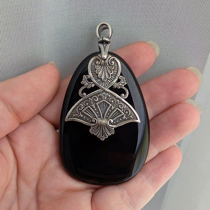 Decorative black druzy stone pendant with silver intricate design held in a hand against a neutral background.  Gothic Egyptian inspired jewelry.