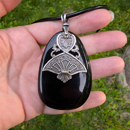 Black druzy stone pendant with silver intricate Egyptian inspired design held in a hand against a blurred green background