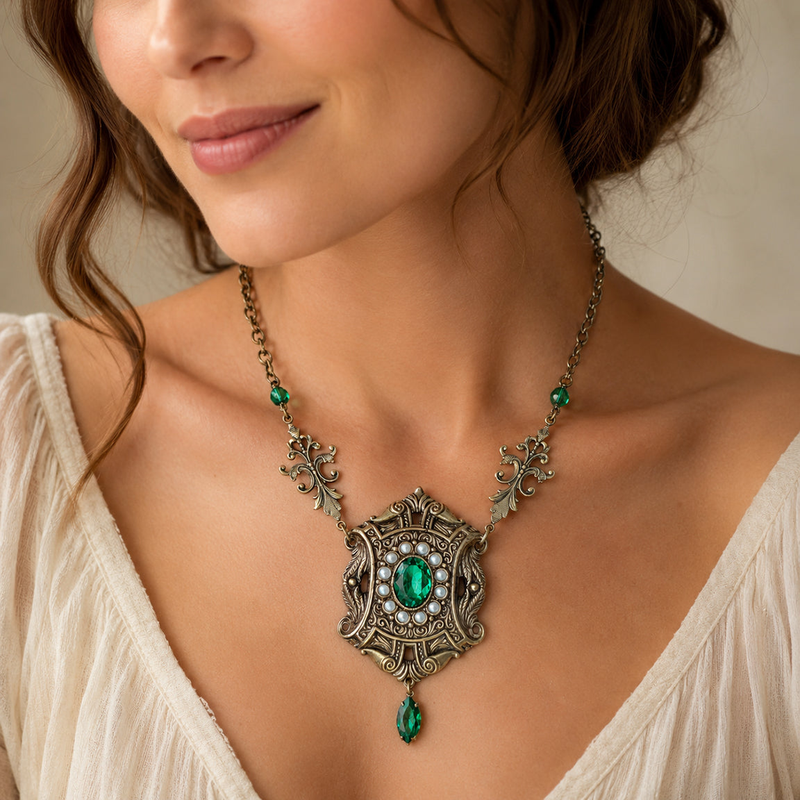 A close-up of a woman wearing an ornate antique-style brass necklace with a large green emerald crystal, surrounded by small cream colored pearls and an Emerald green accent Navette stone.