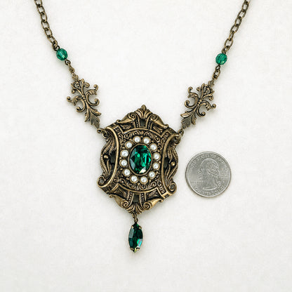 An ornate antique-style antique brass pendant necklace with emerald green crystals and cream pearls, shown next to a US quarter for scale on a white background.