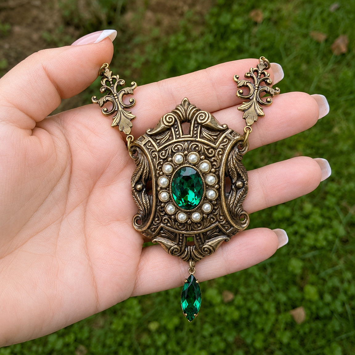 An ornate antique-style brass Victorian pendant necklace with a green emerald center stone, surrounded by cream colored pearls, held in a hand against a blurred green grass background.