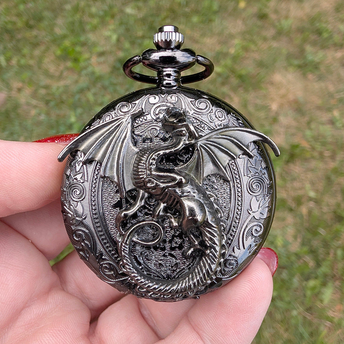 A dark metallic gothic pocket watch pendant featuring an embossed dragon design, held in a person's hand against a blurred green grass background.