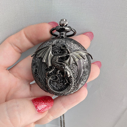 A hand with red glitter nail polish holds a round, dark metallic fantasy pocket watch pendant featuring a detailed dragon design on the front.