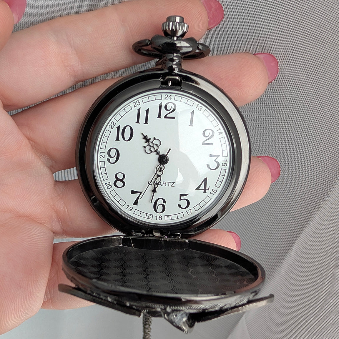 A vintage-style pocket watch with a black metal case and a white dial, held open in a person's hand against a light gray fabric background.