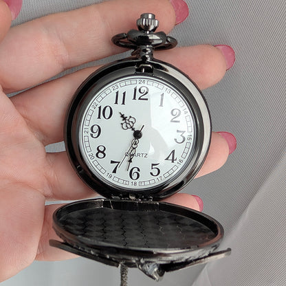 A vintage-style pocket watch with a black metal case and a white dial, held open in a person's hand against a light gray fabric background.