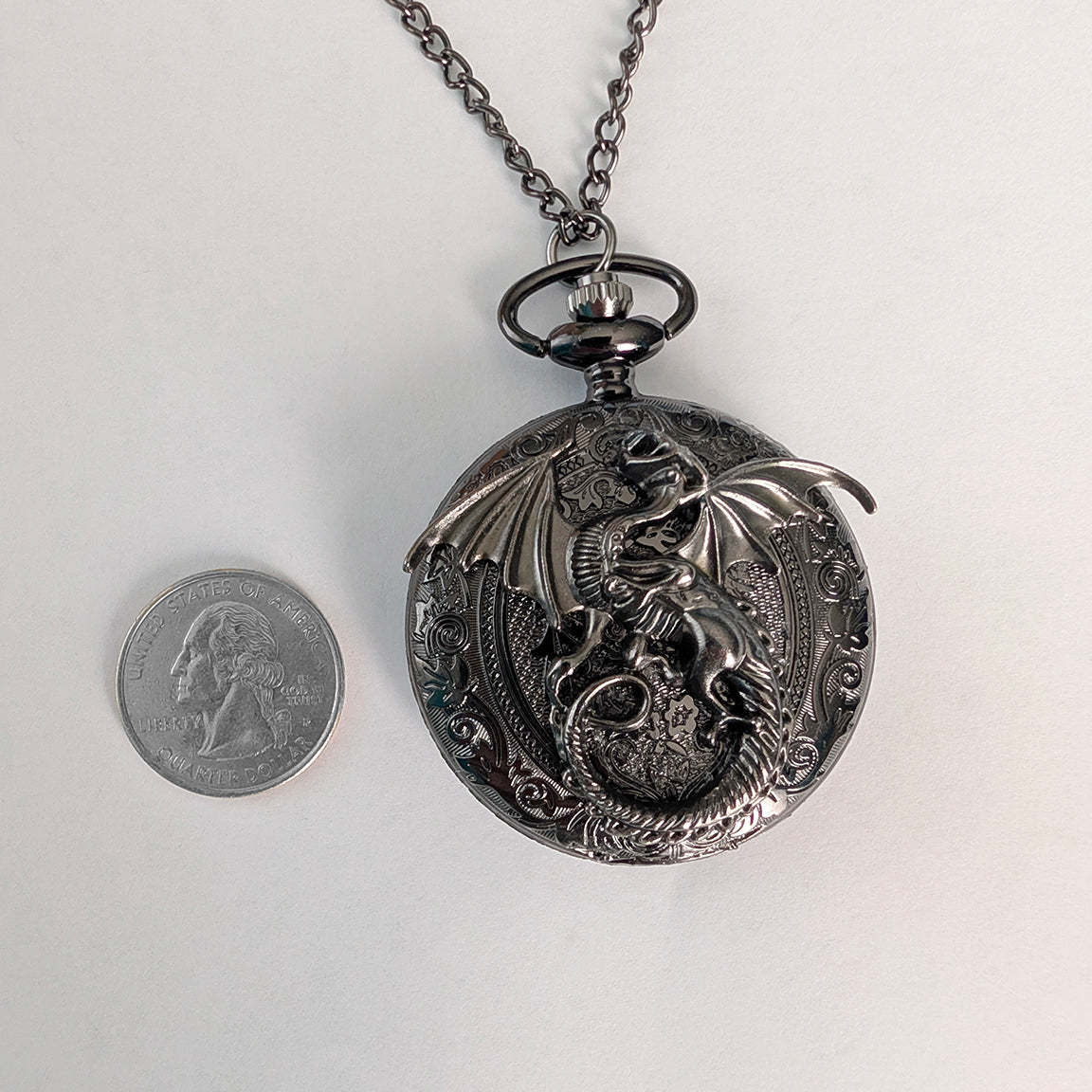 A gunmetal black dragon pocket watch necklace displayed next to a US quarter coin for scale, on a plain white background.