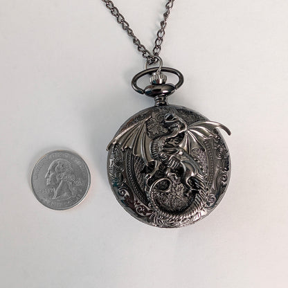 A gunmetal black dragon pocket watch necklace displayed next to a US quarter coin for scale, on a plain white background.