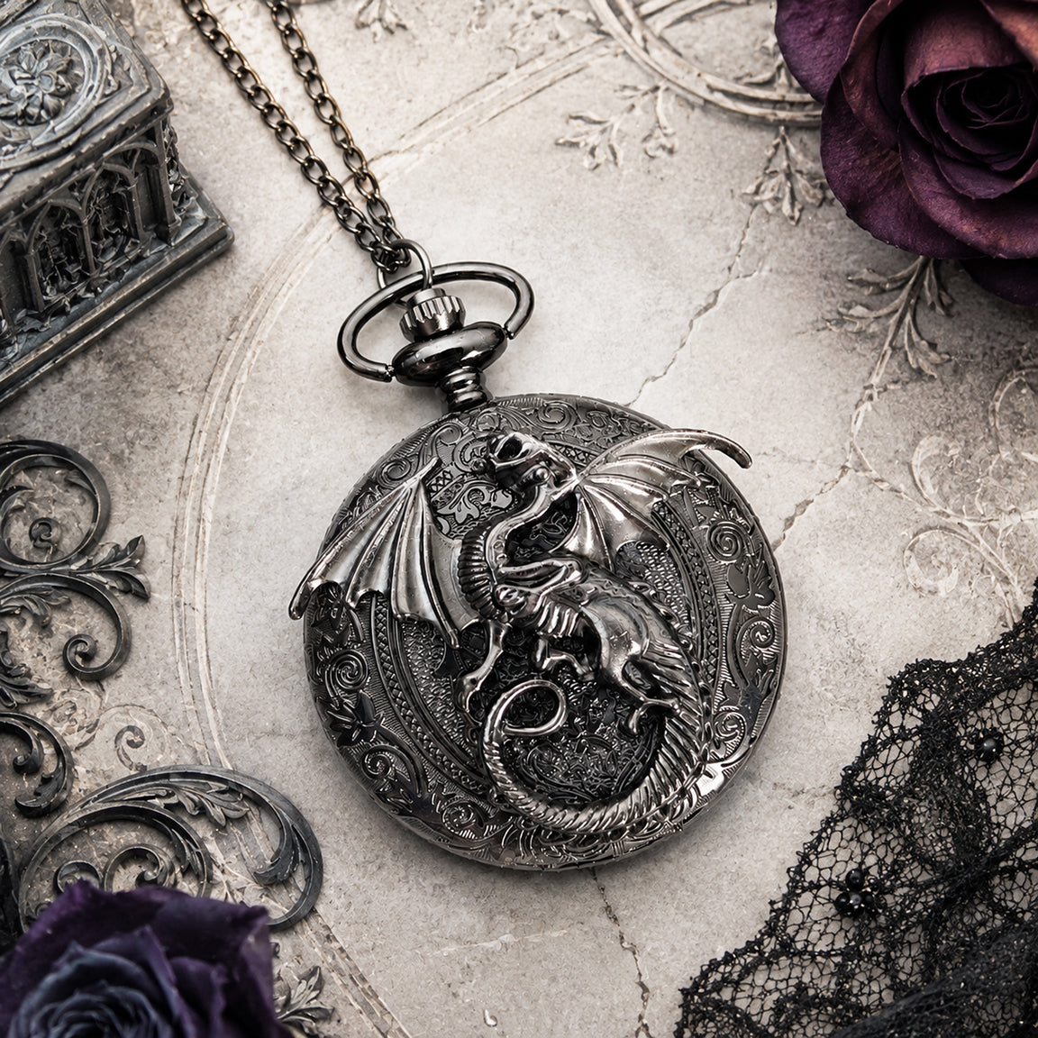 A gothic-style gun metal toned dragon pocket watch necklace displayed on a textured stone surface with vintage props including a rose, lace, and an ornate box.