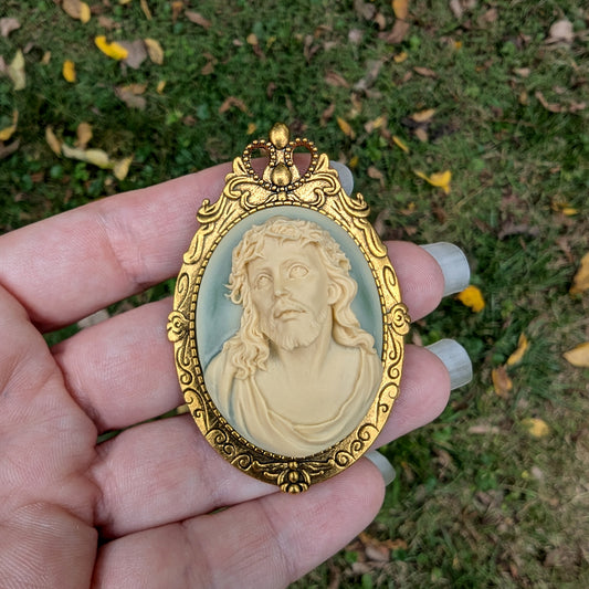 Gold-framed Jesus cameo pendant on a hand against a natural background