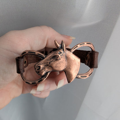 Bronze horse head design on a leather strap bracelet held by a hand against a neutral background
