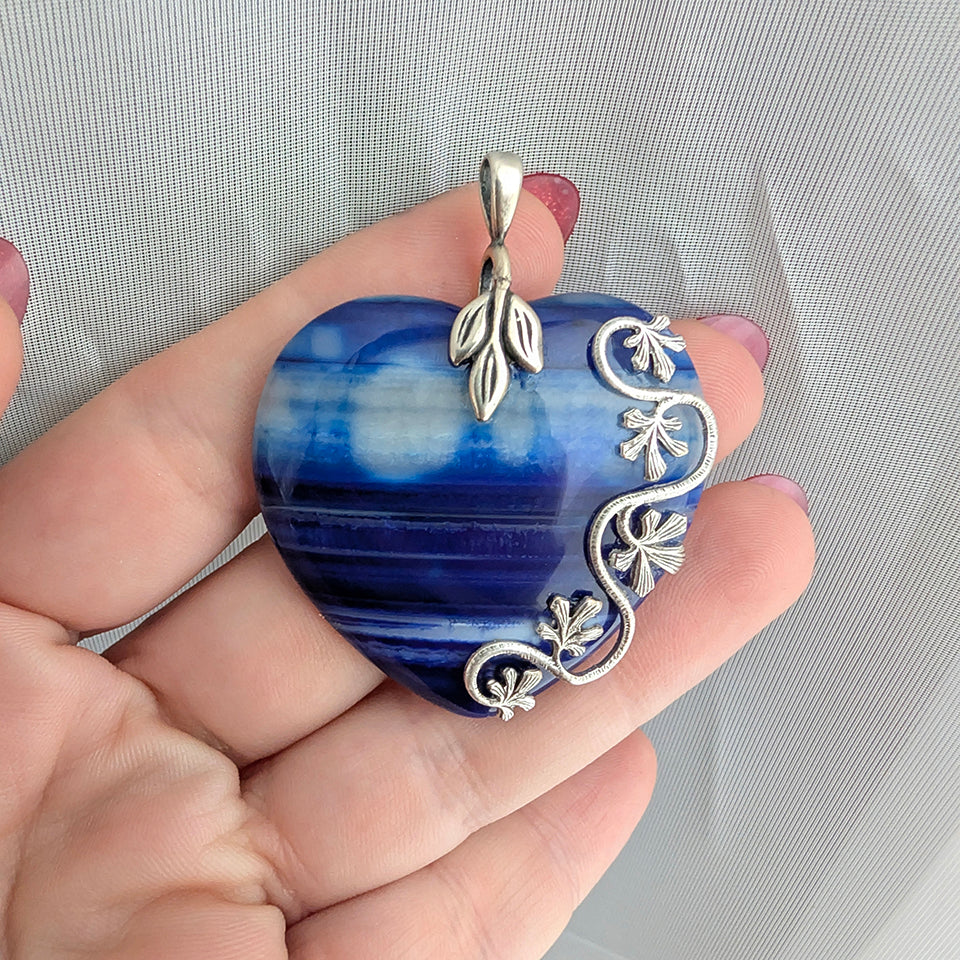 Natural blue striped agate heart pendant featuring sterling silver plated brass accents being held in hand with white background.  Nature Inspired Boho Jewelry.