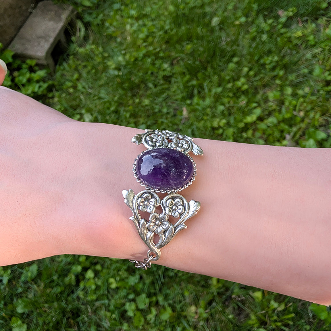 Silver bracelet with a purple gemstone on a wrist against a grassy background.  Vintage Amethyst Bracelet with Floral Detail.  Art Nouveau Sterling Silver Jewelry