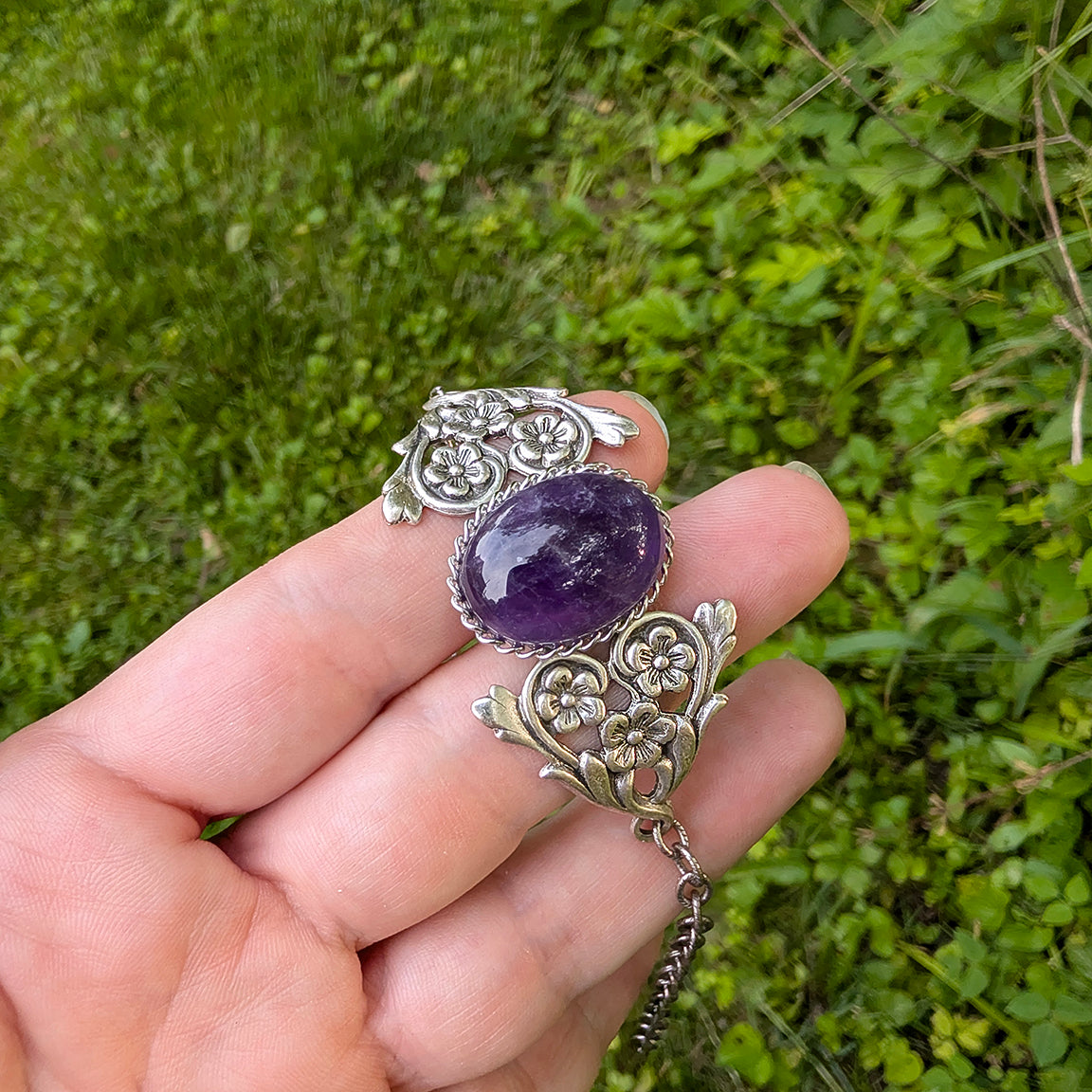 Silver bracelet with a purple gemstone held in a hand against a green grass background.  Vintage Amethyst Bracelet with Floral Detail.  Art Nouveau Sterling Silver Jewelry