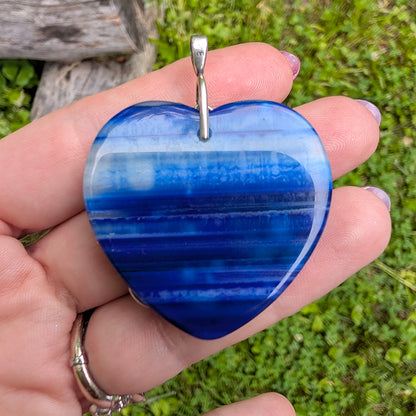 Backside of Natural blue striped agate heart pendant featuring sterling silver plated brass accents being held in hand with grassy background. Nature inspired Boho Jewelry.