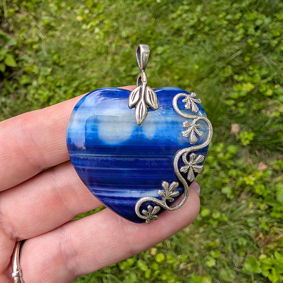 Natural blue striped agate heart pendant featuring sterling silver plated brass accents being held in hand with grassy background. Nature inspired Boho Jewelry.