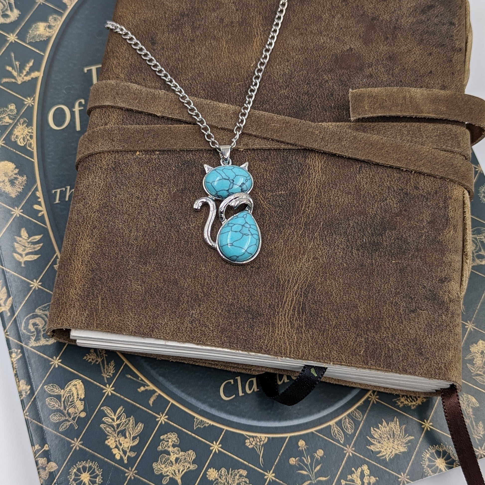 Turquoise cat-shaped pendant on a chain over a brown leather-bound book with decorative cover