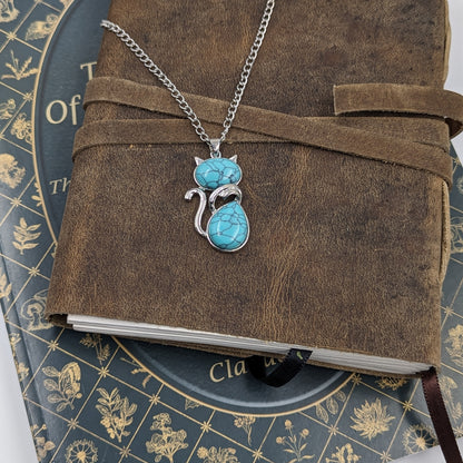 Turquoise cat-shaped pendant on a chain over a brown leather-bound book with decorative cover