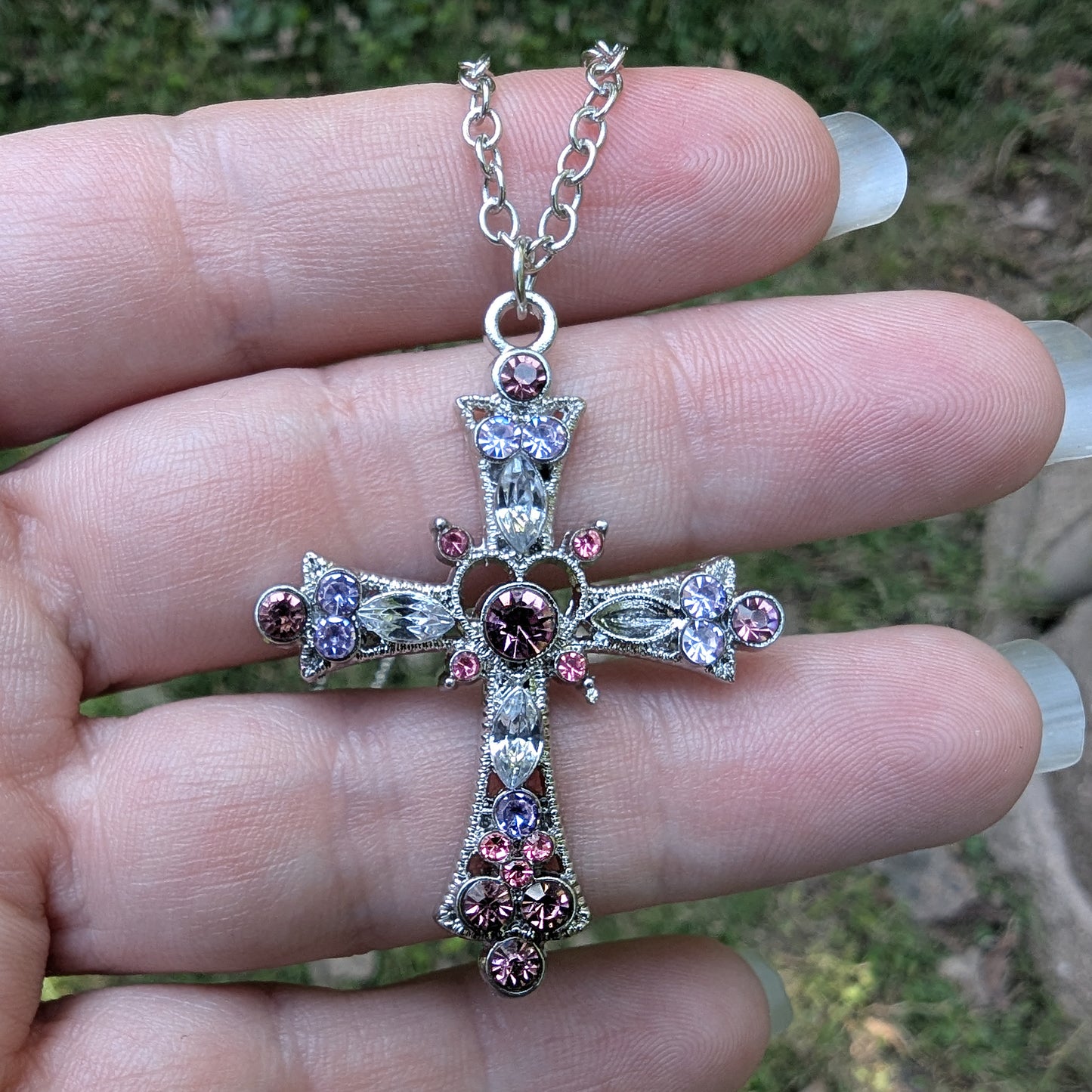 Silver cross necklace with pink and purple gemstones held between fingers against a blurred natural background
