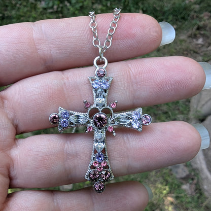 Silver cross necklace with pink and purple gemstones held between fingers against a blurred natural background