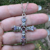 Silver cross necklace with pink and purple gemstones held between fingers against a blurred natural background