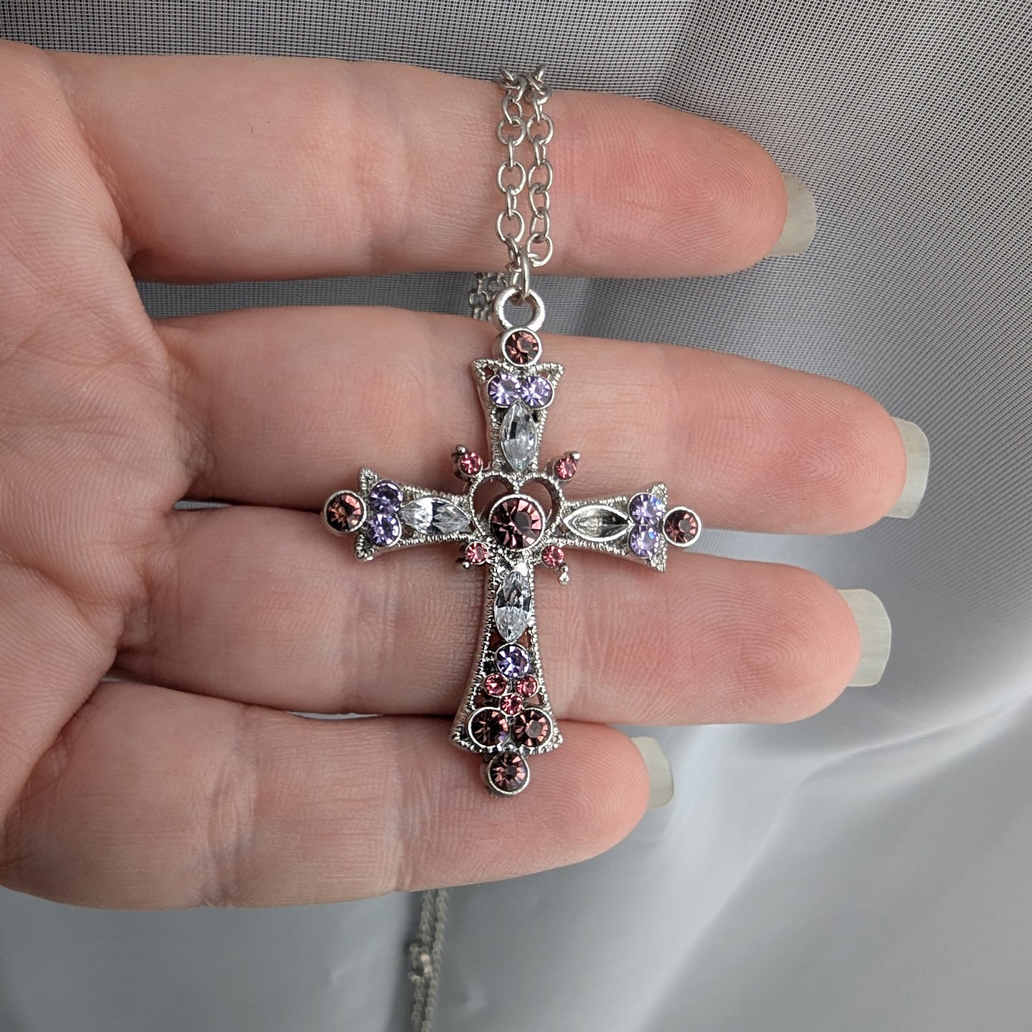 Silver cross pendant with gemstones held between fingers against a neutral background