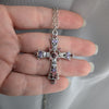 Silver cross pendant with gemstones held between fingers against a neutral background