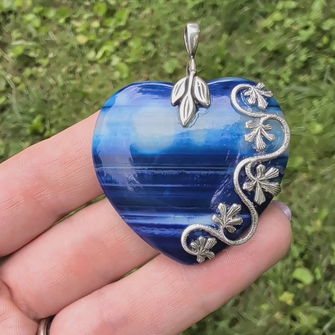 Natural blue striped agate heart pendant featuring sterling silver plated brass accents being held in hand with grassy background. Nature inspired Boho Jewelry.